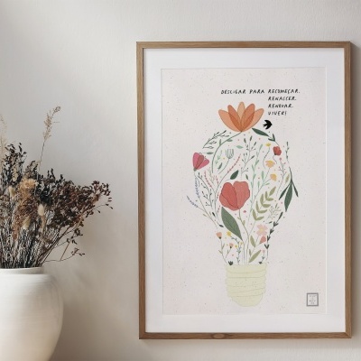 Poster emoldurado com ilustração de lâmpada com flores e texto sobre ideias para economia, na parede ao lado de vaso branco com flores secas