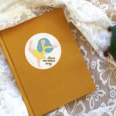 Caderno amarelo com autocolante 'dance YOUR TROUBLES away' sobre tecido rendado branco com figura decorativa de pássaro