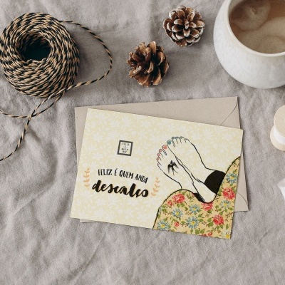 Cartão postal ilustrado com pés femininos e texto 'FELIZ É QUEM AMA descalço', envelope beige, cordão e caneca sobre tecido cinza.