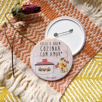 Botão redondo com alfinete e desenho de panela e jarro, texto em português, sobre tecido amarelo e manta laranja com flor seca