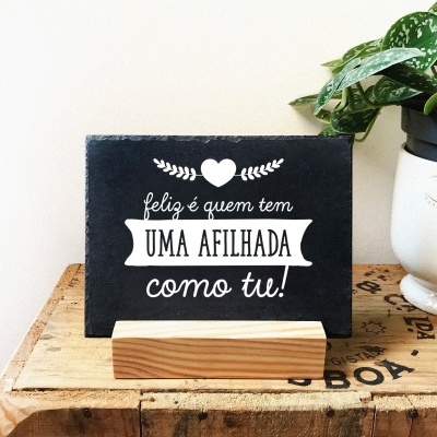 Placa preta com mensagem para afilhada em suporte de madeira ao lado de vaso branco com planta
