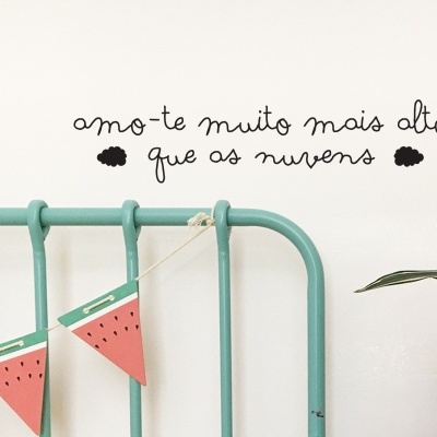 Texto decorativo em português na parede branca acima de uma cabeceira de cama verde água com guirlanda de bandeirolas de melancia