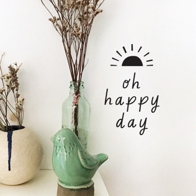 Decoração com vaso creme, garrafa verde, estátua de pássaro e vinil na parede com texto 'oh happy day'.