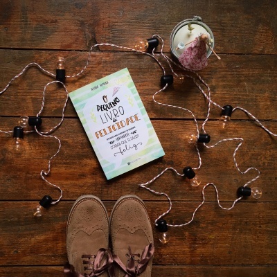 Livro 'Pequeno Livro da Felicidade' sobre chão de madeira com luzes decorativas e sapatos castanhos