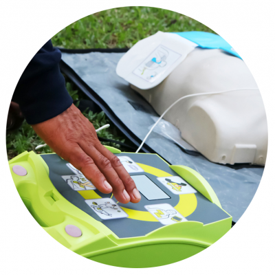 Desfibrilhador automático verde e cinza com mão a usar, manequim de treino ao fundo