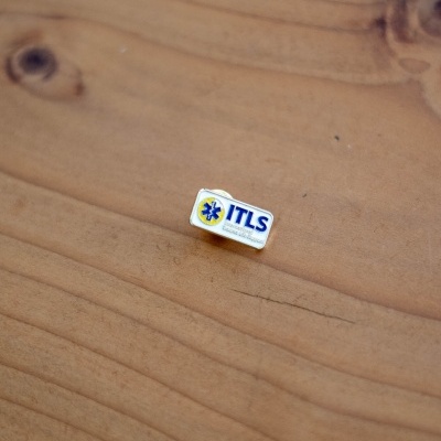 Pin metálico branco e amarelo com texto ITLS sobre madeira clara