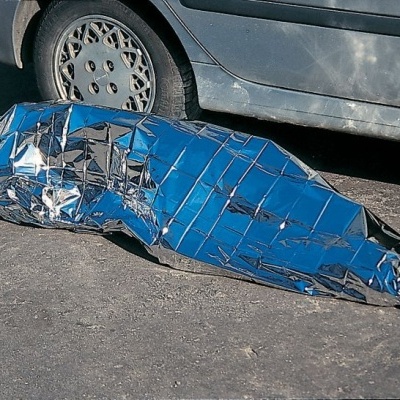 Pessoa enrolada em cobertor isotérmico prateado no chão junto a carro cinzento