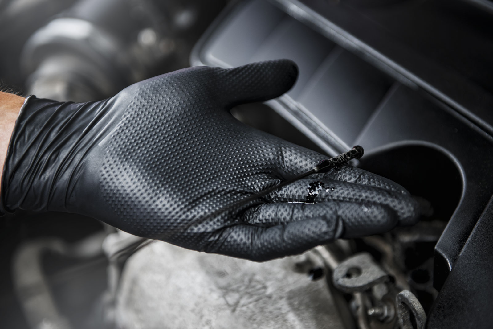 MERCATOR - LUVAS GOGRIP TEXTURIZADAS PRETAS E LARANJA (CX 50 Luvas) Mão com luva preta segurando vareta de óleo em motor de carro