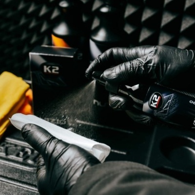 Frasco de limpeza automóvel K2 com mãos com luvas pretas e panos amarelos no fundo