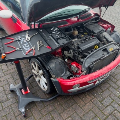Carro vermelho com capô aberto e suporte de ferramentas preto ao lado.