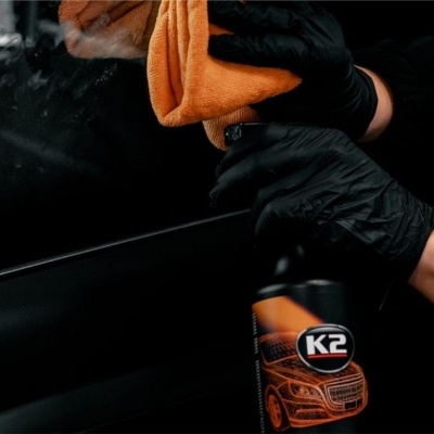 Mão com luva preta a limpar superfície preta brilhante com pano laranja e frasco K2
