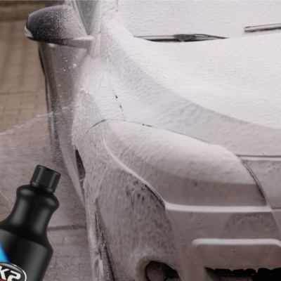 Carro cinzento coberto de espuma branca com produto de limpeza K2 em frasco preto e azul em cenário exterior pavimentado
