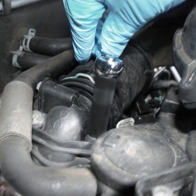 Mão com luva azul segurando ferramenta no motor de carro com mangueiras pretas e peças metálicas