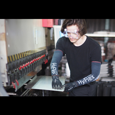 Trabalhador operando prensa metálica com luvas HyFlex e mangas de proteção, em ambiente industrial