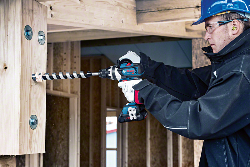 Homem com capacete azul e óculos de proteção perfura madeira com furadeira em construção