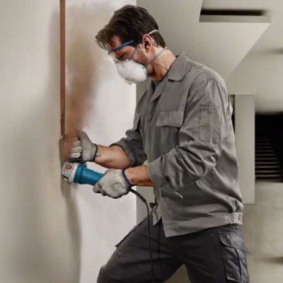 Homem com máscara e óculos a lixar parede branca com lixadeira elétrica.