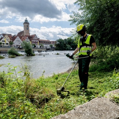 Trabalhador com colete refletor e capacete usa aparador de relva à beira de um rio
