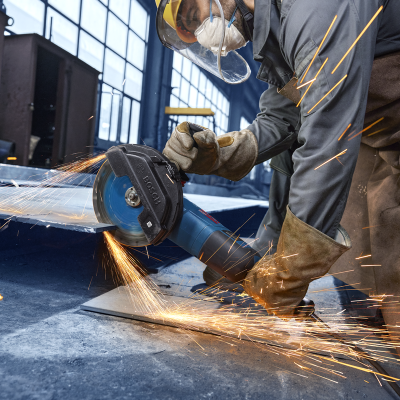 Trabalhador em ambiente industrial usando rebarbadora Bosch azul e equipamento de proteção a cortar chapa metálica com faíscas.