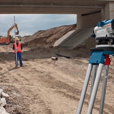 Operários de construção usam equipamento topográfico em terreno arenoso com escavadora ao fundo