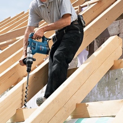 Homem a furar vigas de madeira com furadeira elétrica azul numa construção ao ar livre