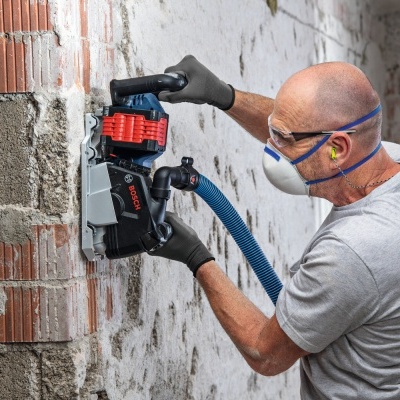 Homem a usar lixadora Bosch elétrica numa parede de tijolos com equipamento de proteção