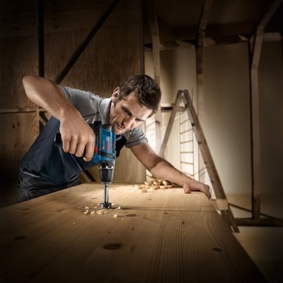 Homem a usar furadeira elétrica azul numa tábua de madeira clara em espaço de trabalho incompleto