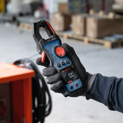 Multímetro de pinça Bosch azul e preto com mostrador digital segurado por mão com luva preta.