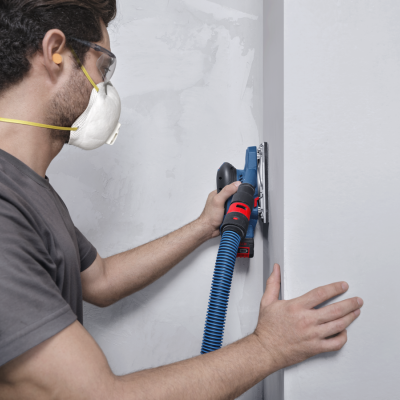 Homem com máscara a usar lixadora elétrica azul numa parede