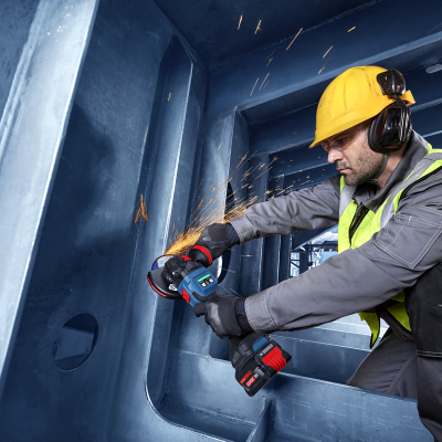 Trabalhador com capacete e colete a usar rebarbadora elétrica em estrutura metálica azul
