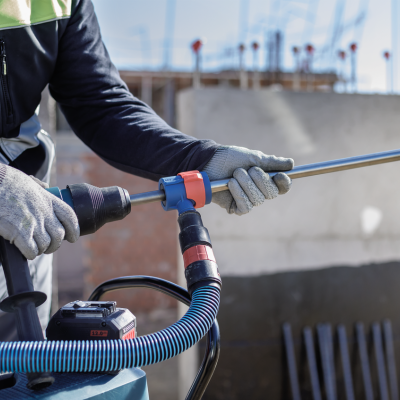 Pessoa usando ferramenta elétrica Bosch azul e preta em obra com parede cinza e estrutura de ferro ao fundo