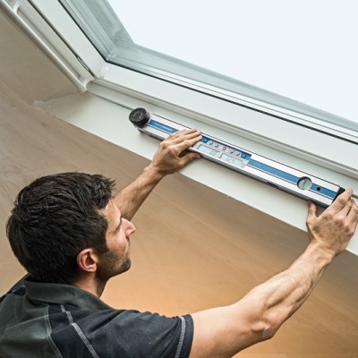 Homem a usar nível de bolha para medir janela numa divisão com paredes claras e teto inclinado