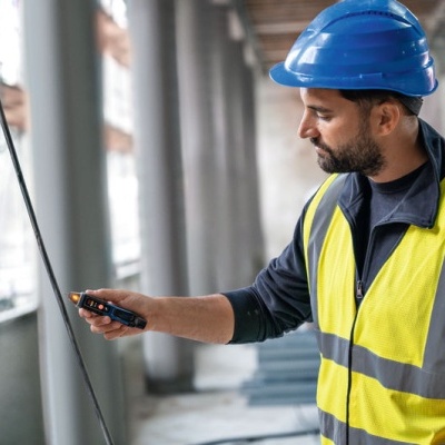 Trabalhador de construção com capacete azul e colete refletor amarelo usando aparelho portátil num local de construção