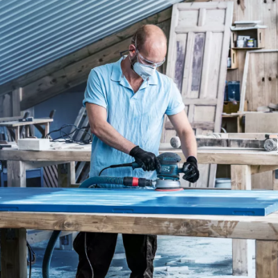 Homem a lixar porta azul em oficina de carpintaria com máscara e luvas