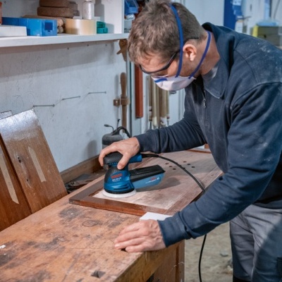 Homem usando lixadeira elétrica para madeira numa oficina com portas de madeira ao fundo