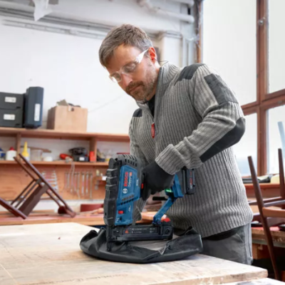 Homem a usar ferramenta elétrica Bosch numa oficina com madeira e ferramentas.