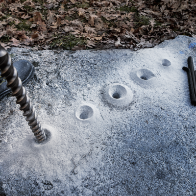 Pedra cinzenta com buracos circulares, broca espiral e marcador preto ao lado, chão com folhas
