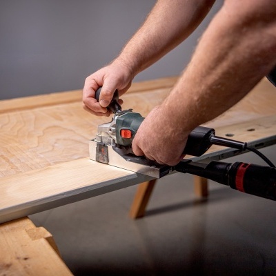 Pessoa lixa a borda de uma peça de madeira clara com lixadora elétrica preta e cinza