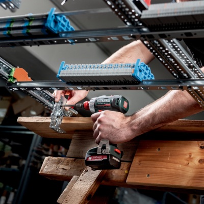 Homem usando furadeira para montar estrutura metálica numa bancada de madeira numa oficina