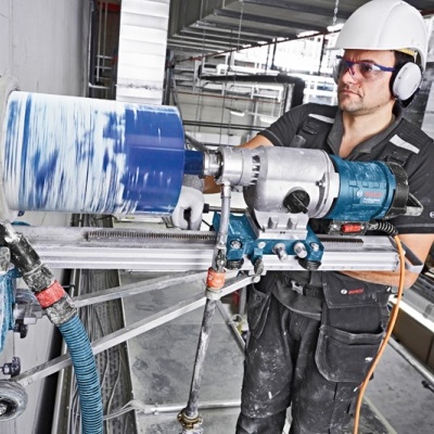 Homem com capacete, óculos e protetores auriculares a perfurar parede com broca industrial
