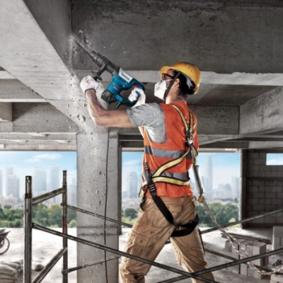 Trabalhador da construção civil com capacete e colete laranja usando furadeira em edifício de concreto inacabado