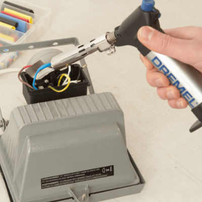 Mãos seguram isqueiro Dremel perto de estação de soldagem com acessórios coloridos.