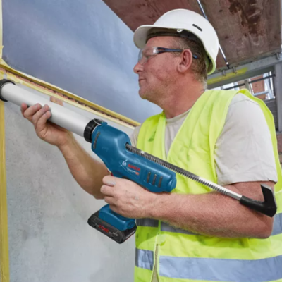 Homem com colete refletor e capacete aplicando material branco na parede com ferramenta azul.