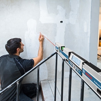 Homem a medir uma parede branca com fita métrica num espaço interior com escada e corrimão preto.