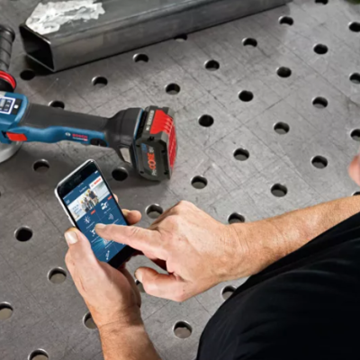 Esmerilhadora azul e preta com bateria vermelha sobre mesa metálica perfurada e pessoa a usar smartphone com app Bosch.