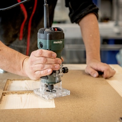 Pessoa usando tupia elétrica verde e preta metabo a cortar madeira numa oficina