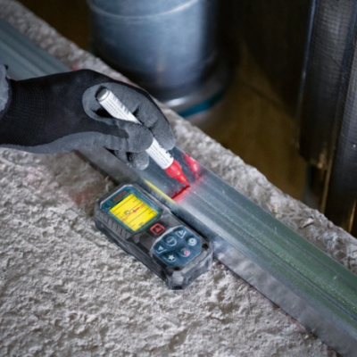 Medidor laser digital preto sobre superfície de construção com barra metálica e mão com luva preta apontando dispositivo