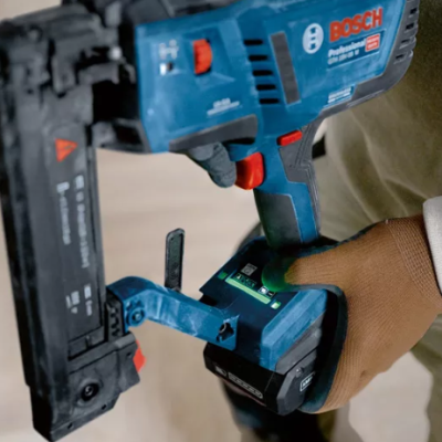 Pistola de pregos Bosch azul e preta segurada por pessoa com luvas marrons em fundo de madeira