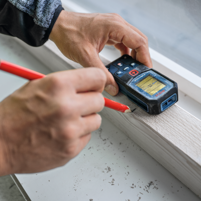 Mãos usando lápis vermelho e medidor laser Bosch numa superfície de madeira clara