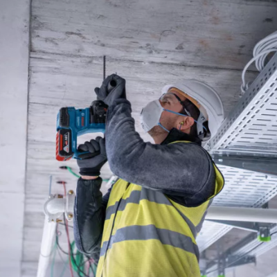 Trabalhador com capacete e colete refletor usando furadeira azul no teto da obra