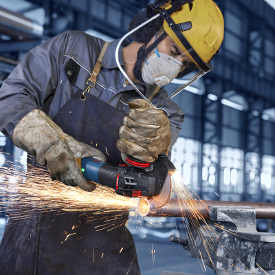 Trabalhador em fábrica usa esmeriladora para cortar tubo metálico com equipamentos de proteção.